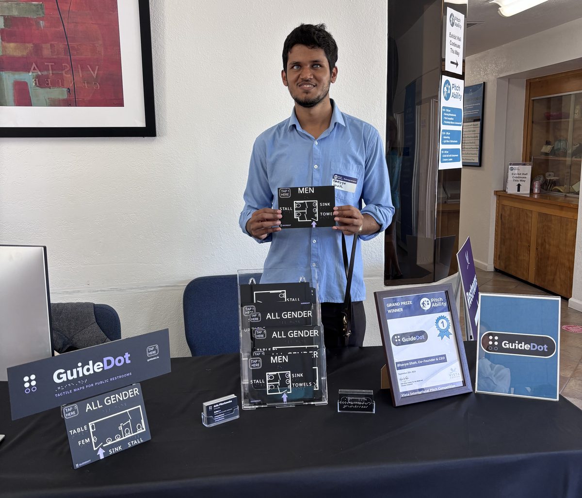 Bhavya smiles behind the GuideDot table at Vista Center in San Jose, holding up a small black MEN map with a TAP HERE pad, white raised linework and the labels STALL, SINK and TOWELS. On the black tablecloth in front of him: a GuideDot sign, a stand of ALL GENDER and MEN maps, an ALL GENDER map on an easel, a braille business card, and a framed Grand Prize Winner certificate from the PitchAbility competition.