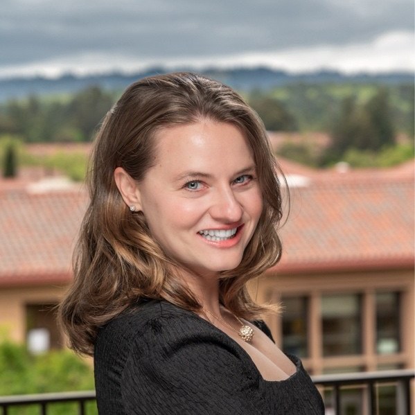 Ania Filochowska, a young woman with shoulder-length light brown hair and a wide smile, in a black long-sleeved top with her arms crossed, photographed outdoors on a balcony with terracotta rooftops, trees and an overcast sky behind her.