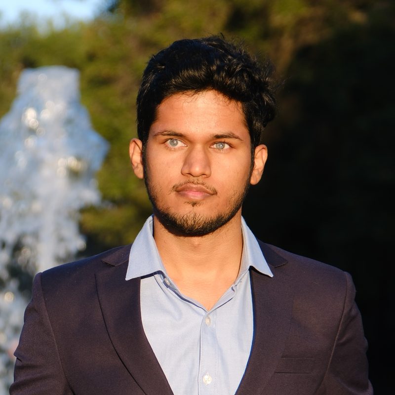 Bhavya Shah, a young man with short dark hair and a light beard, in a navy blazer over a blue open-collared shirt, photographed outdoors in warm evening light with a fountain and trees behind him.